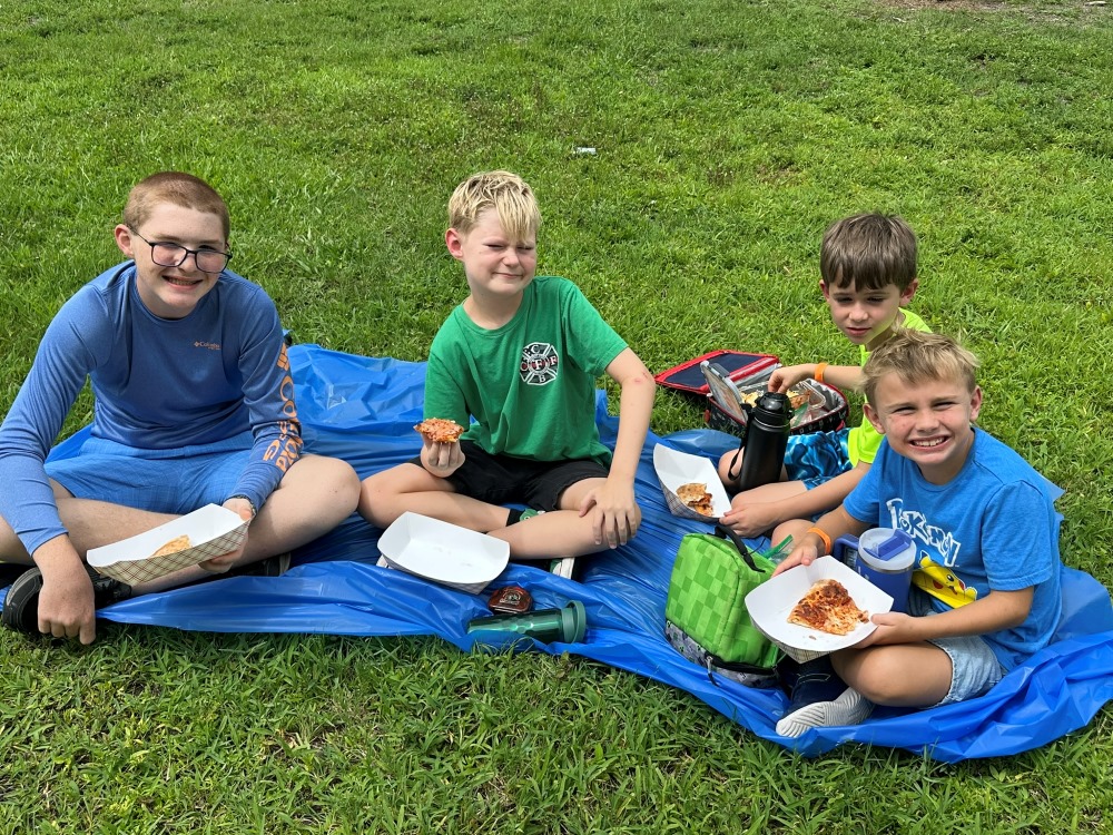 Four boys sit on a blue blanket in the grass, eating slices of pizza